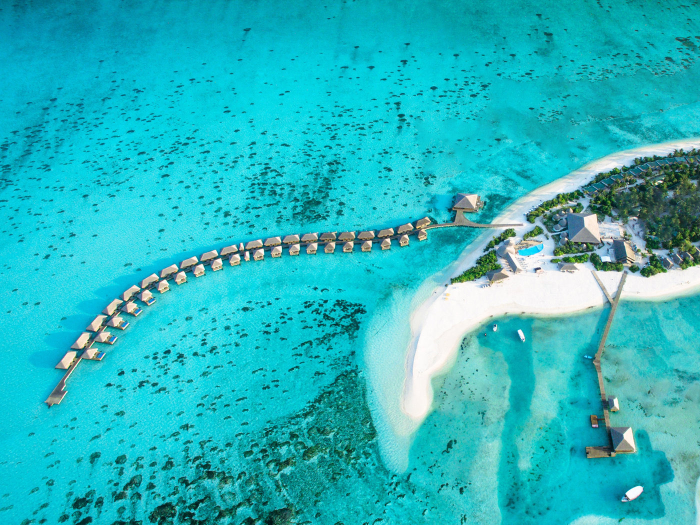 Aerial view of the resort's overwater villa row curving away from a bright white sandbank and the island's palm-covered tip.