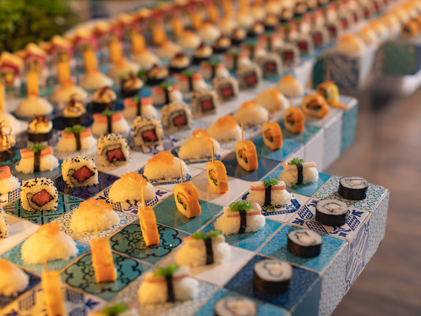 A long buffet counter of patterned blue and white tiles carrying dozens of individually plated sushi and maki pieces.
