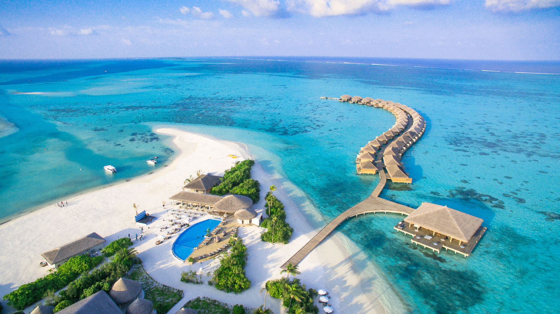 Aerial view of Cocoon Maldives: a curving arc of thatched overwater villas reaching into a turquoise lagoon, with the island's pool, restaurants and white sand beach in the foreground.