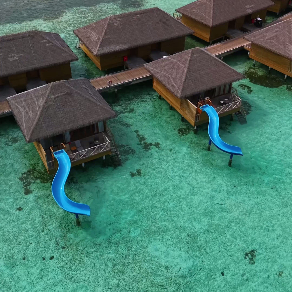 A row of thatched overwater villas linked by a timber walkway, two of them with blue slides descending into clear green water.