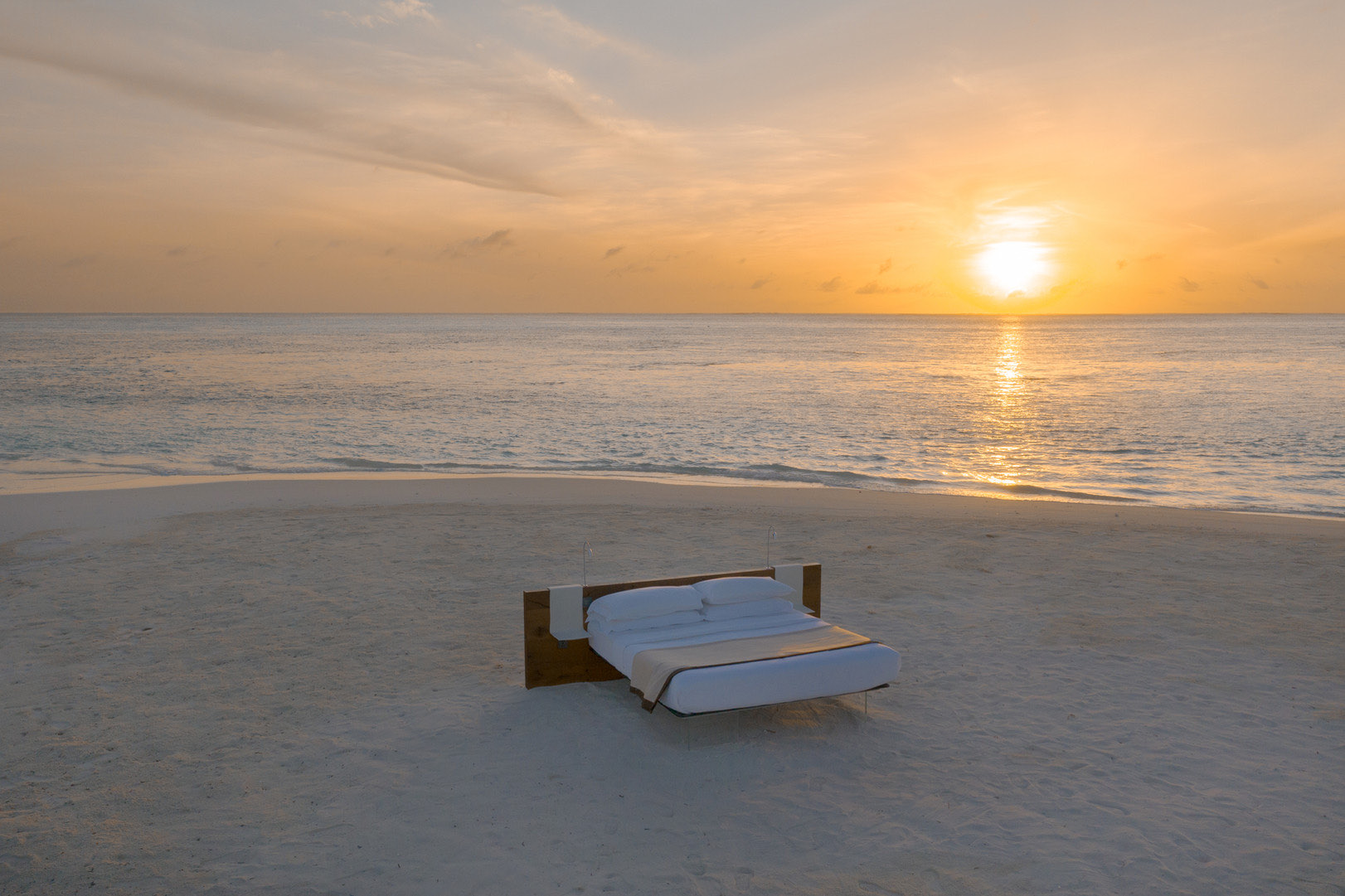 A dressed daybed standing alone on empty white sand as the sun sets low over a calm ocean, the sky washed gold.