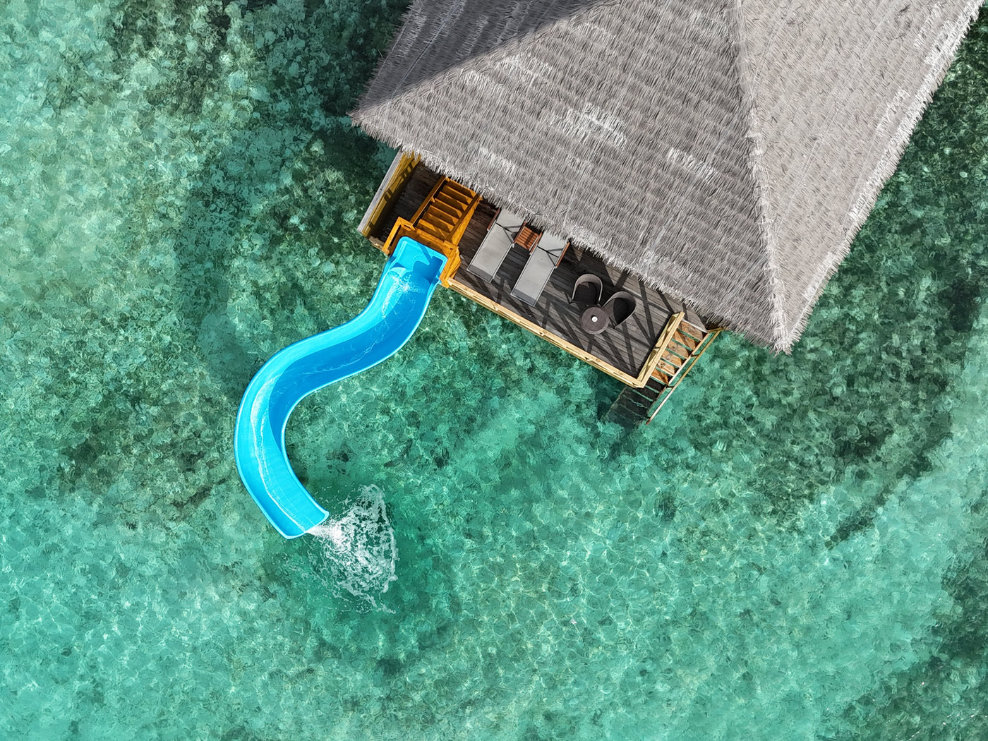 Overhead view of an overwater villa with a bright blue water slide curving from its deck into the shallow turquoise lagoon.